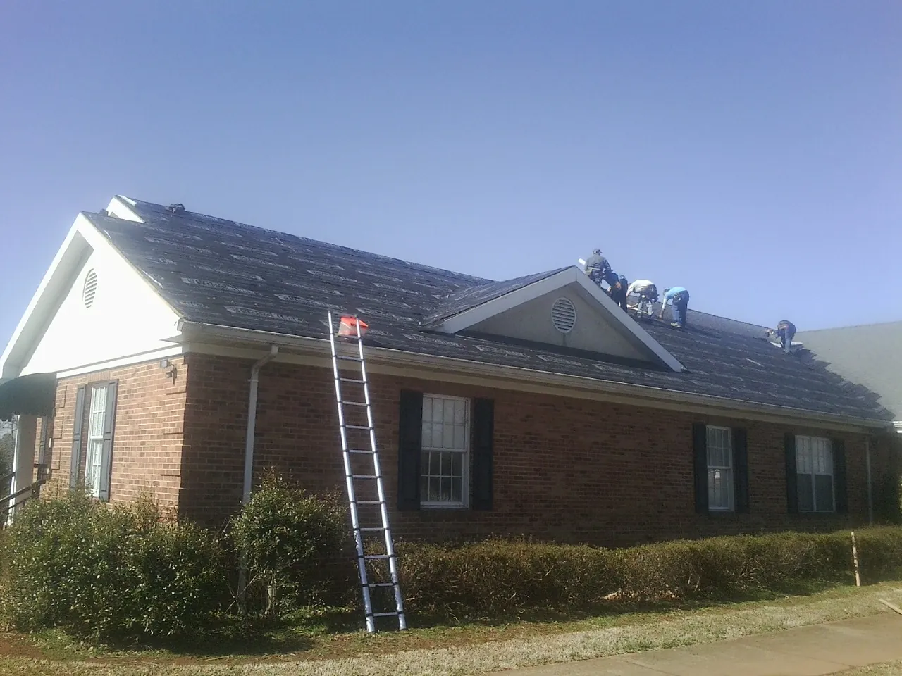 Crew tearing shingles off a brick ranch roof with a ladder set — Metal Roof Repair work by our crew in Whitmire