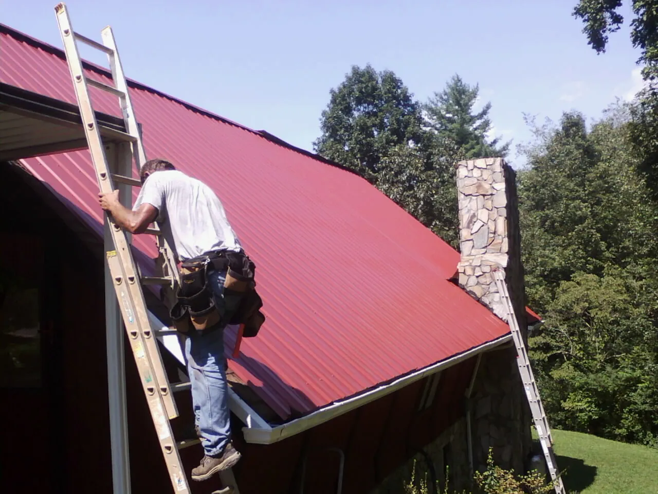Roofer on a ladder beside a red metal roof and stone chimney — Soffit & Fascia Repair by Roofing Brothers in Whitmire